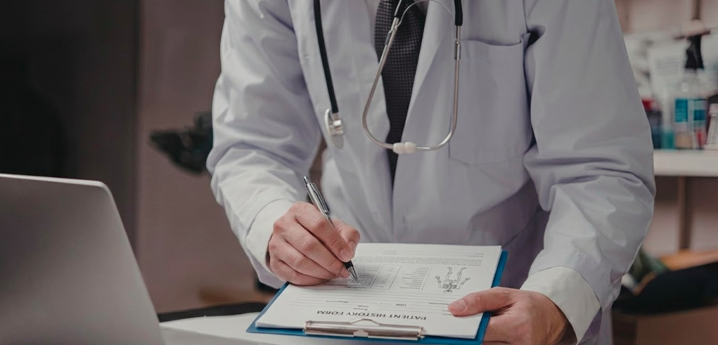 A physician writing notes next to a laptop, the kind of consistent publishing that makes surgeons findable by AI